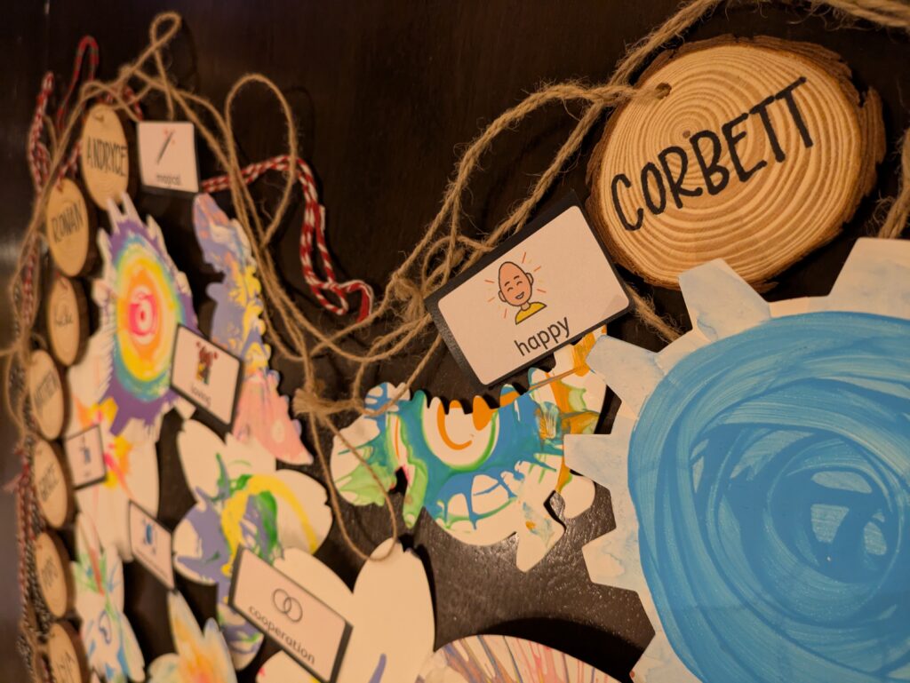 PXL_20250518_035234562 (2) Close up of Happy Heart, with a picture communication symbol of the word happy and a wood cookie name tag with the name Corbett.